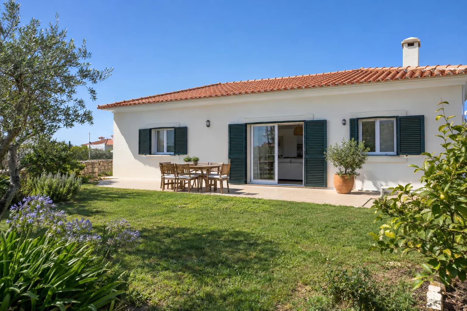 Moradia de um piso com fachada de reboco claro, telhado de telha cerâmica e um pátio com mesa exterior.